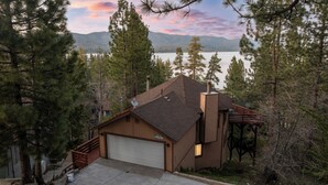 A two-story house with a garage, surrounded by tall pine trees and a lake view. - New: Luxury Lakeview Cabin, Deck, Fireplace, View (Big Bear Lake)