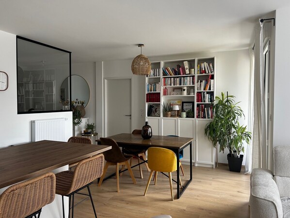 A dining area with a wooden table, wicker chairs, and a yellow chair. There is a bookshelf filled with books and decorative items.