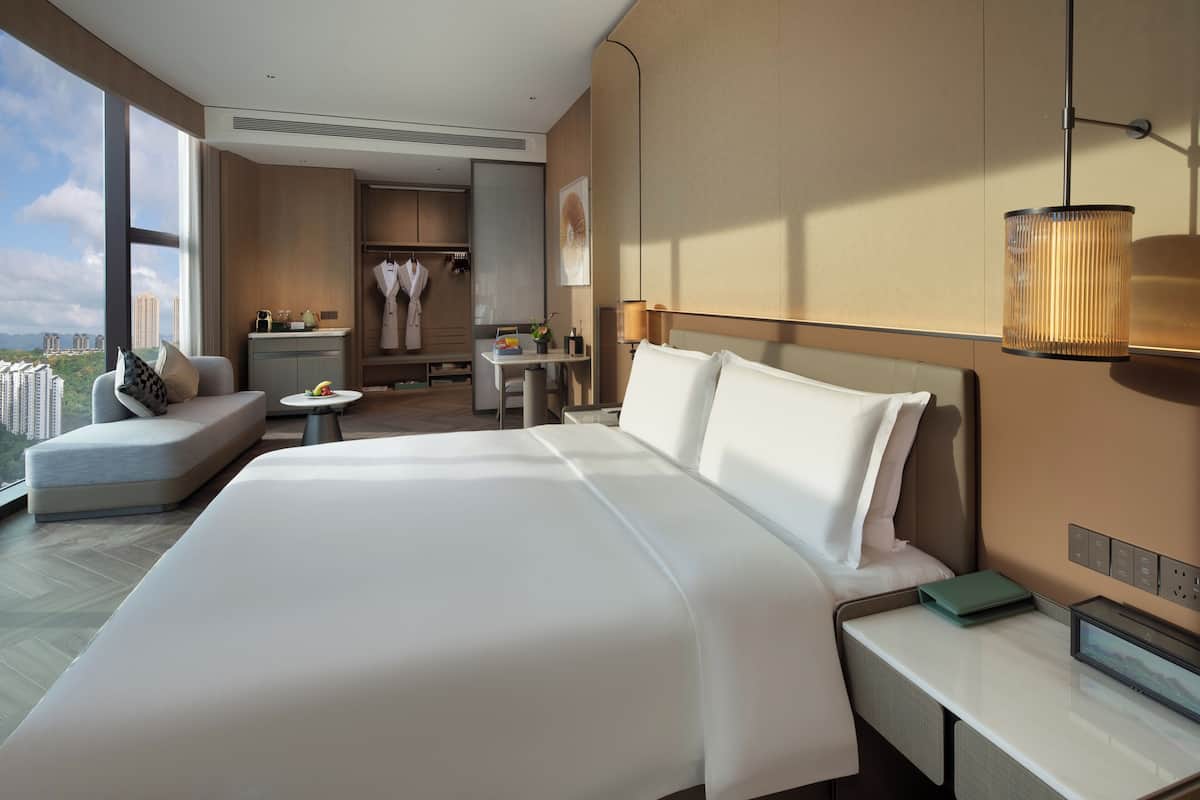 A modern hotel room with a large bed, a desk, a seating area, and a view of the cityscape.