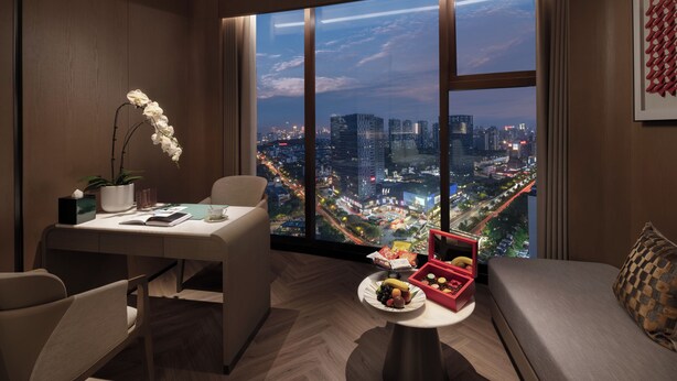 Una habitación de hotel moderna con un ventanal grande que ofrece vistas a la ciudad, una zona de comedor con mesa y sillas, y un pequeño rincón de descanso con un sofá y una mesita auxiliar.