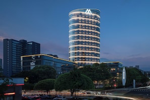 A modern high-rise building with a distinctive curved design and illuminated windows, surrounded by trees and other buildings.