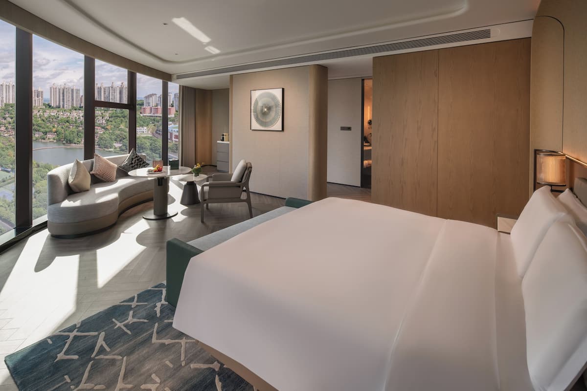 A modern hotel room with a large bed, a curved sofa, a small table, and a view of a cityscape through large windows.