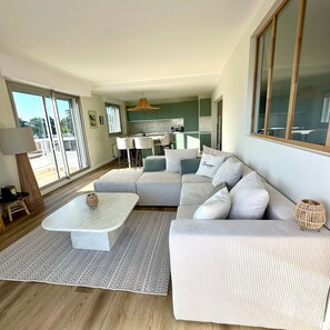 A modern living room with a sectional sofa, a coffee table, and a dining area with a kitchen in the background. - Le Pivoine : vue mer à La Baule (La Baule-Escoublac)