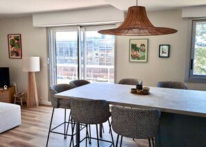 A modern dining area with a large island table, six upholstered chairs, a woven pendant light, and large windows offering a view of the cityscape. - Le Pivoine : vue mer à La Baule (La Baule-Escoublac)