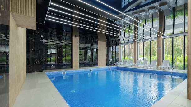 An indoor swimming pool with clear glass walls, modern lighting, and white lounge chairs. - Comfortable Apartment | Pool | Close to the Beach (Pobierowo)