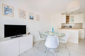A modern kitchen with white cabinets, a dining area with a glass table and white chairs, and a flat-screen TV on a white cabinet. - Le Joli Baulois - La Baule (La Baule-Escoublac)