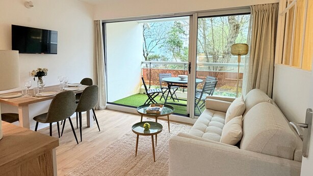 Uma sala de estar moderna com um sofá, uma mesa de jantar posta para quatro pessoas e uma varanda com mesa e cadeiras.