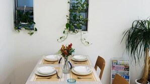 A dining area with a long table set for four, two windows with plants, and a potted plant to the side. - Takara House (Tokyo)