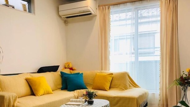 A living room with a yellow sofa, a small round table with a floral tablecloth, and a window with curtains. - Takara House (Tokyo)