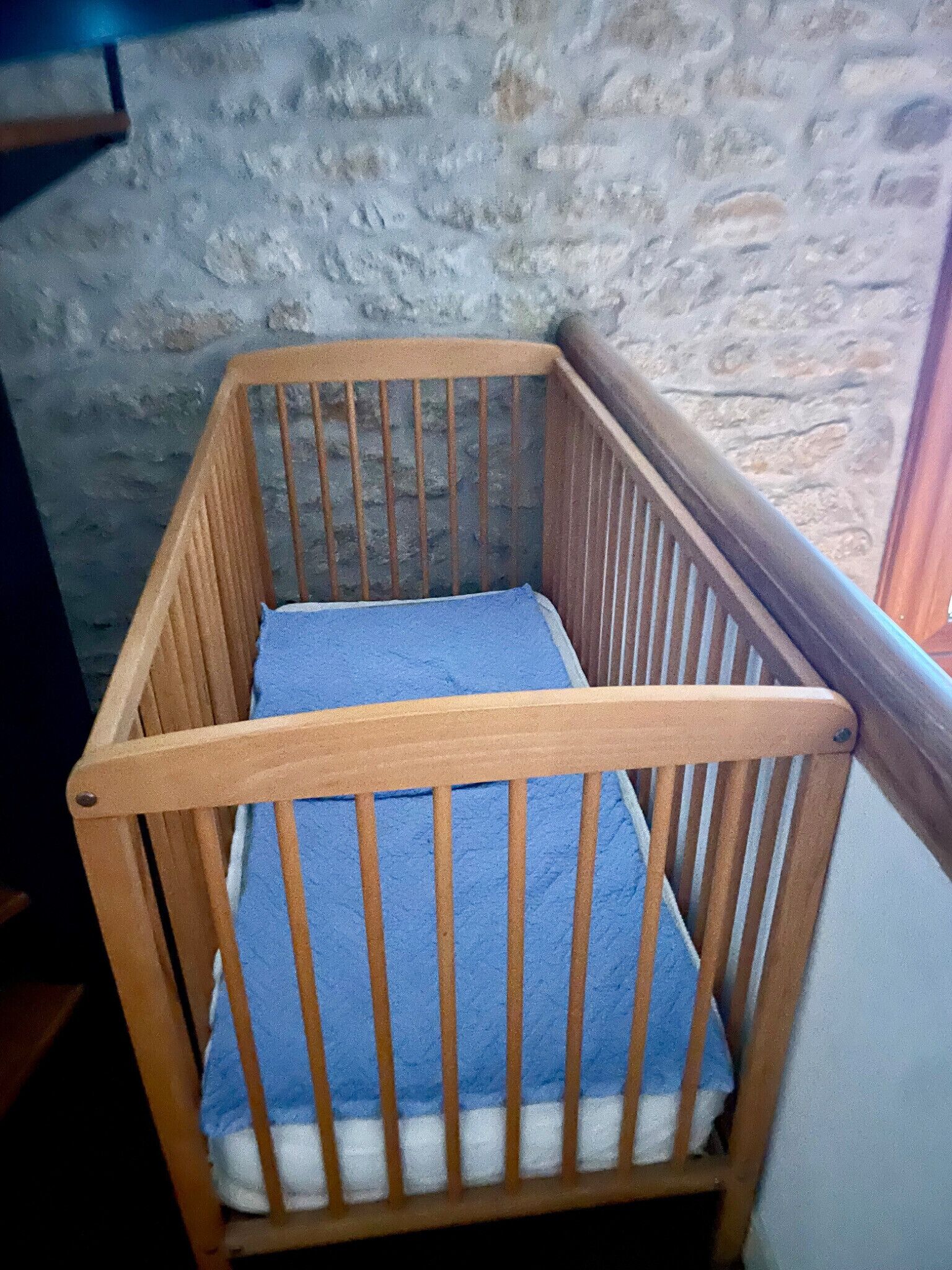 A wooden crib with a blue mattress against a stone wall.