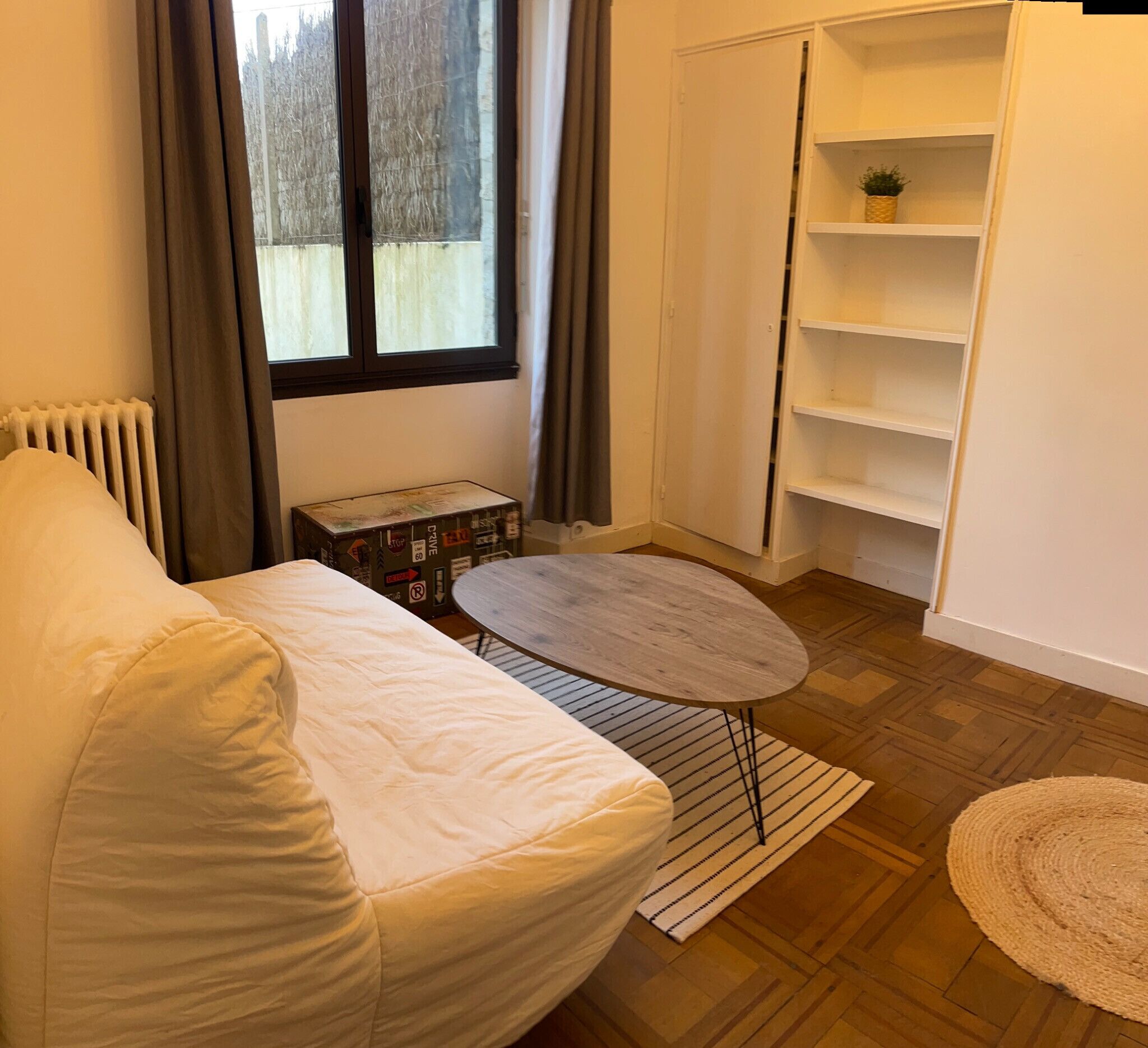 A room with a white sofa, a small table, a radiator, a window with curtains, and a white shelving unit.