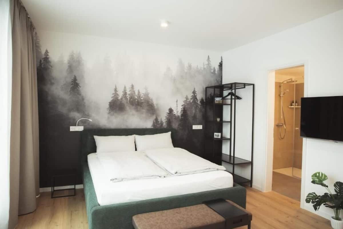 Ein Schlafzimmer mit Bett, Nachttisch, Fernseher, Duschbereich und einer Wand mit Waldmotiv.