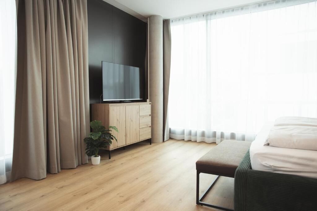 Une chambre d’hôtel moderne avec un plancher en bois, un lit avec une literie blanche, un téléviseur posé sur une commode en bois et une plante en pot.