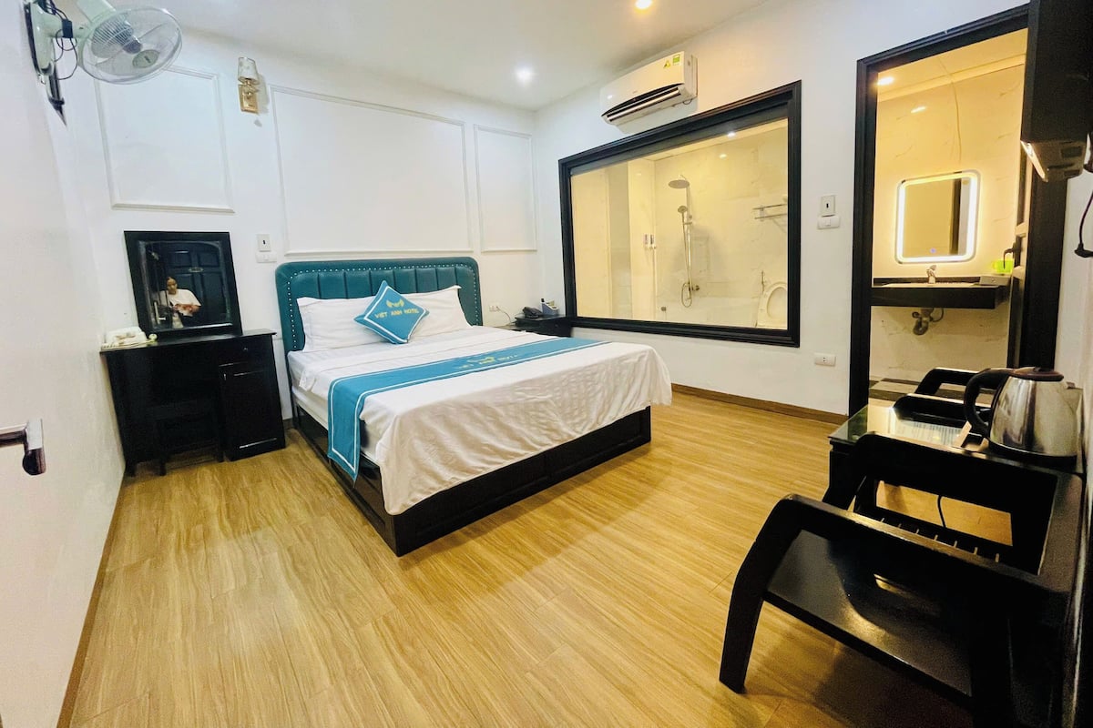 A hotel room with a bed, bedside table, a mirror, a window, and a shower area.