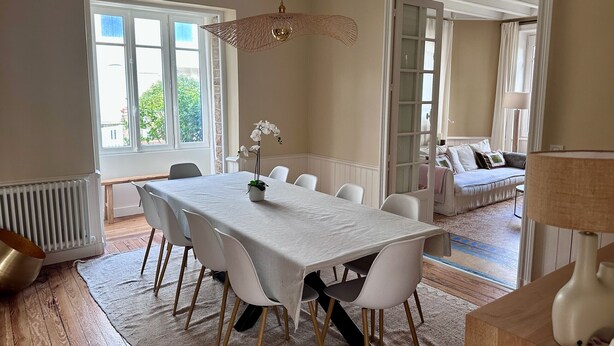A dining area with a white table, chairs, a hanging light fixture, and a view through an open door to a living space.