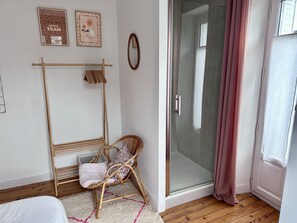 Ein Schlafzimmer mit Bett, Stuhl, Wäscheständer, Duschabtrennung aus Glas und einem Fenster.