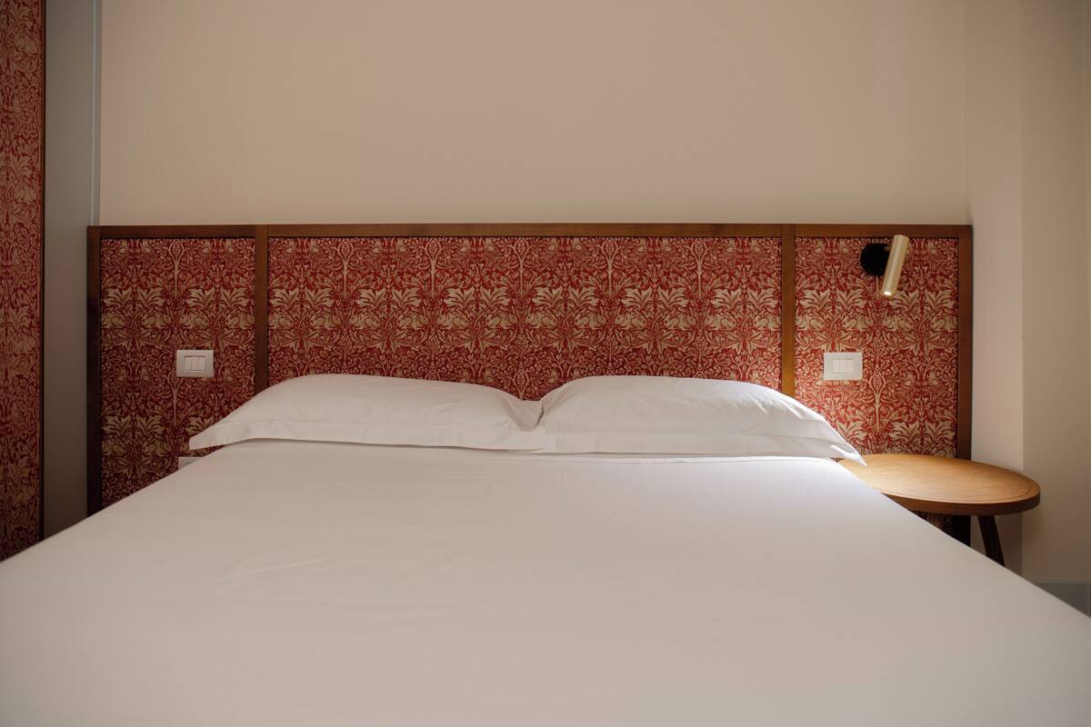 A bed with white linens, a wooden headboard with a floral pattern, a small round table, and a wall-mounted light fixture.