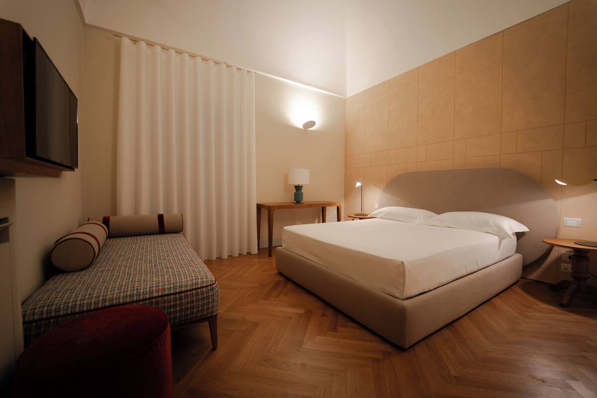 A hotel room with a bed, a small table, a chair, a TV, and a window with curtains.