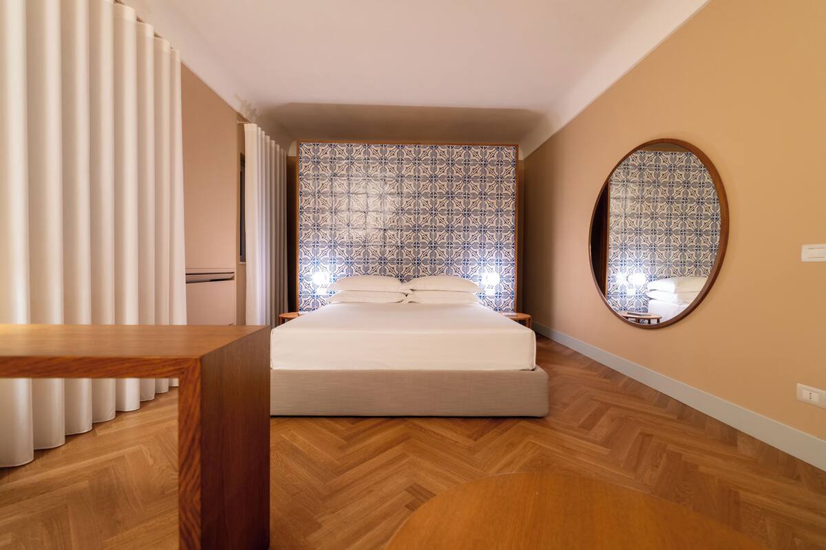 A bedroom with a bed, wooden floor, curtains, a mirror, and patterned wallpaper.