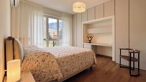 A bedroom with a bed, a desk, a chair, a lamp, and a view of mountains through the window. - Appartamento Belvedere - Affitti Brevi Italia (Lecco)