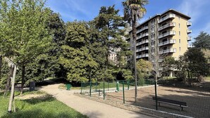 A park with a pathway, trees, and a multi-story building with balconies. - Appartamento Belvedere - Affitti Brevi Italia (Lecco)