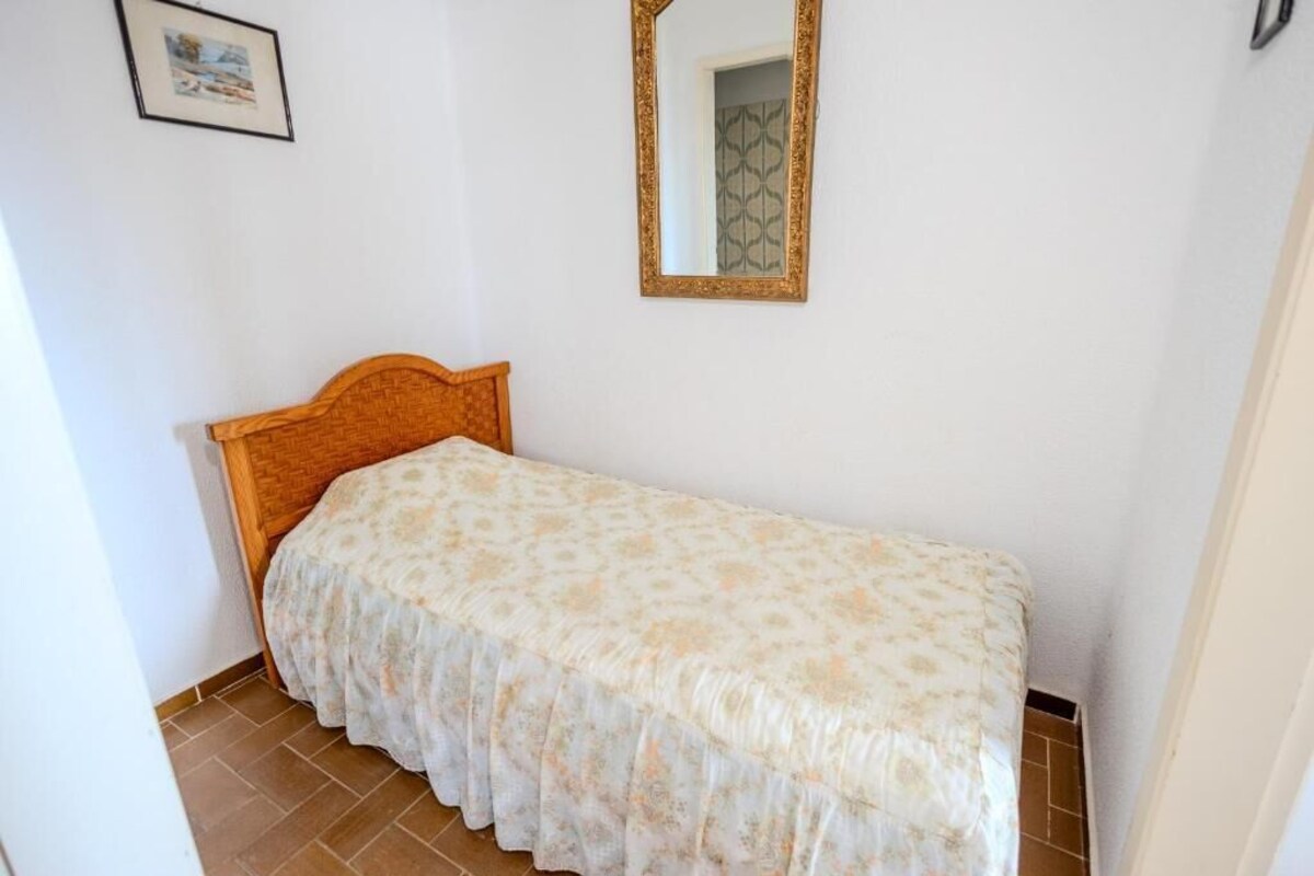 A small, single-bed room with a wooden headboard, a patterned bedspread, a wall-mounted mirror, and a framed picture.
