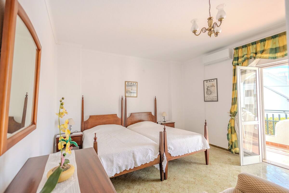Una habitación con dos camas de madera, un espejo, una lámpara de araña y un ventanal con vistas.