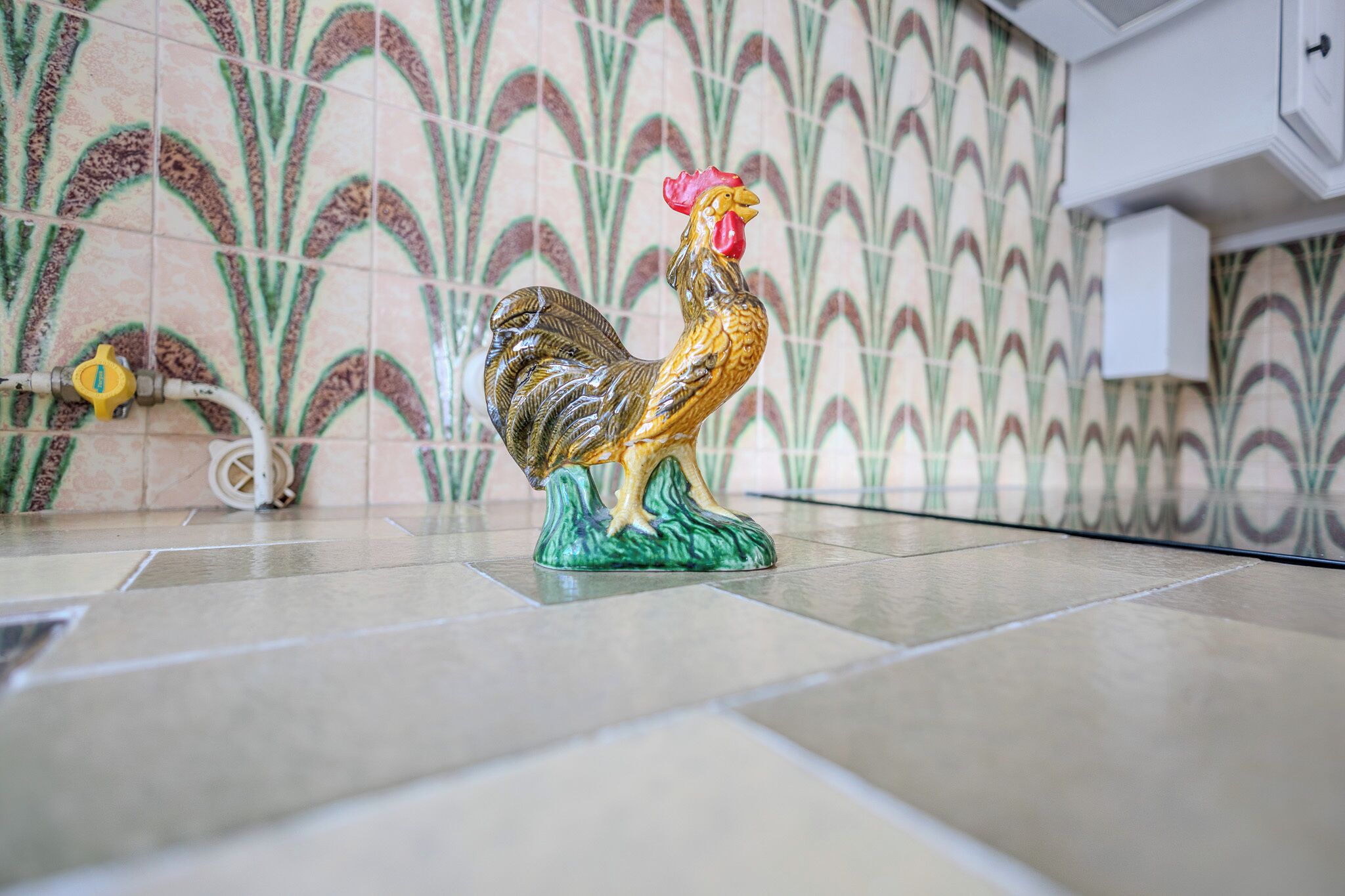Un gallo in ceramica su un pavimento piastrellato, con una parete decorata sullo sfondo.