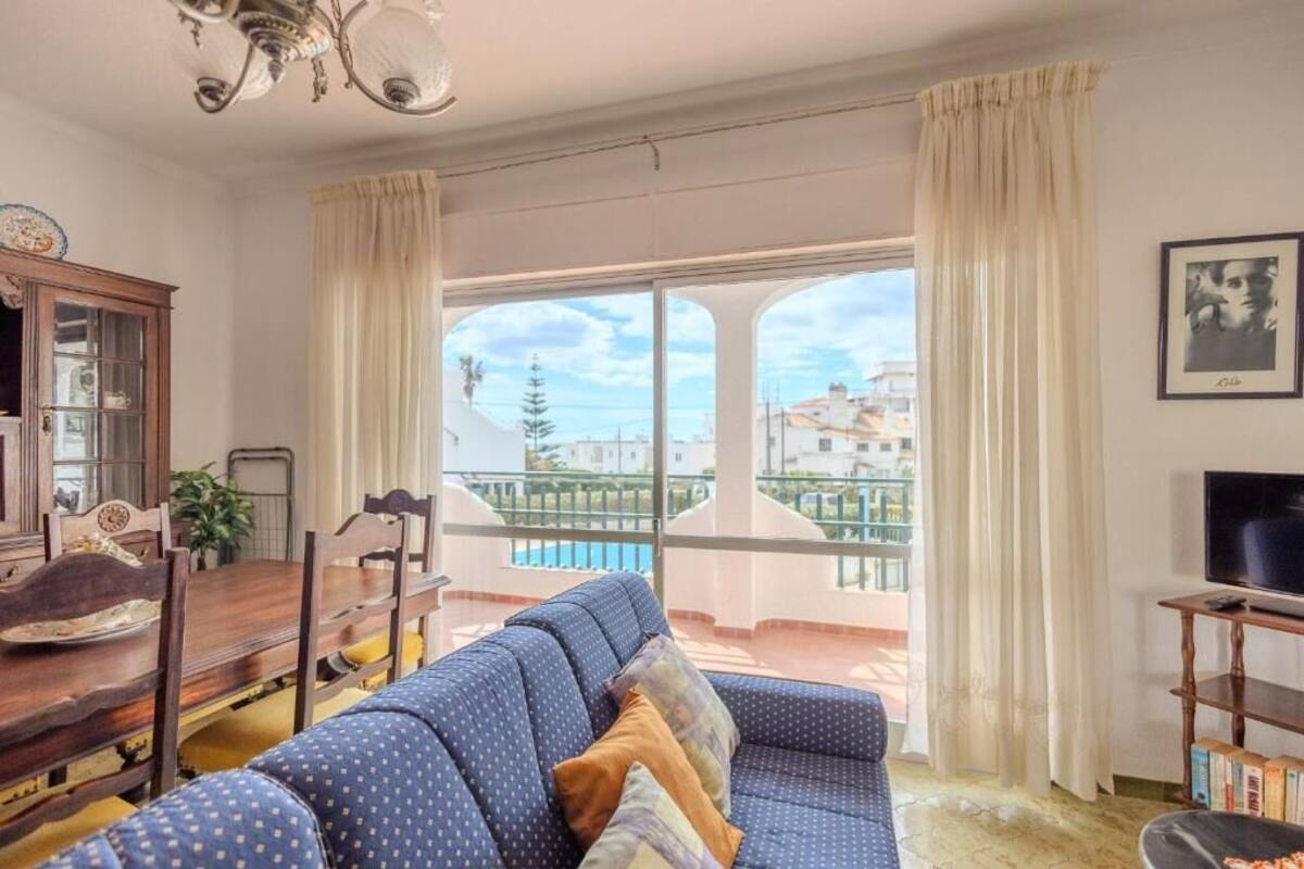 Una sala de estar con un sofá, un televisor, una mesa de comedor y vistas a un balcón con piscina.