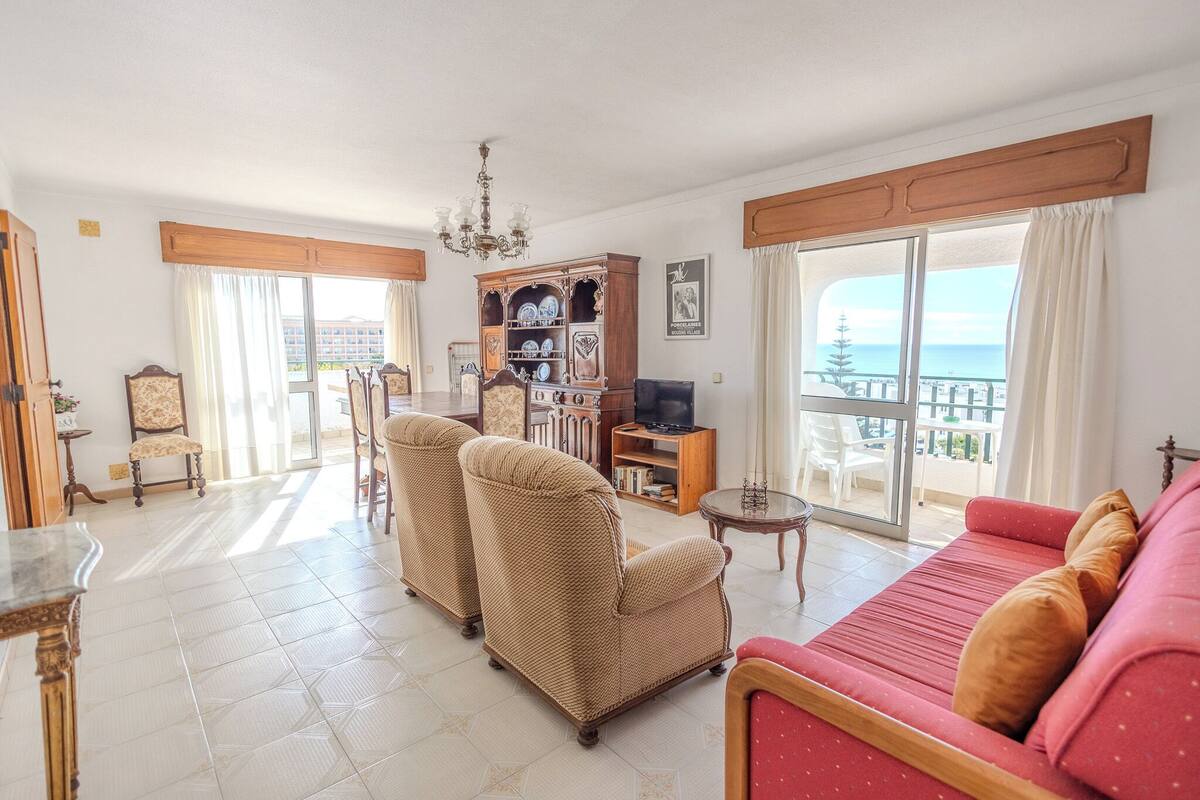 Una sala de estar con sofá, sillas, televisor, estantería y un balcón con vistas.