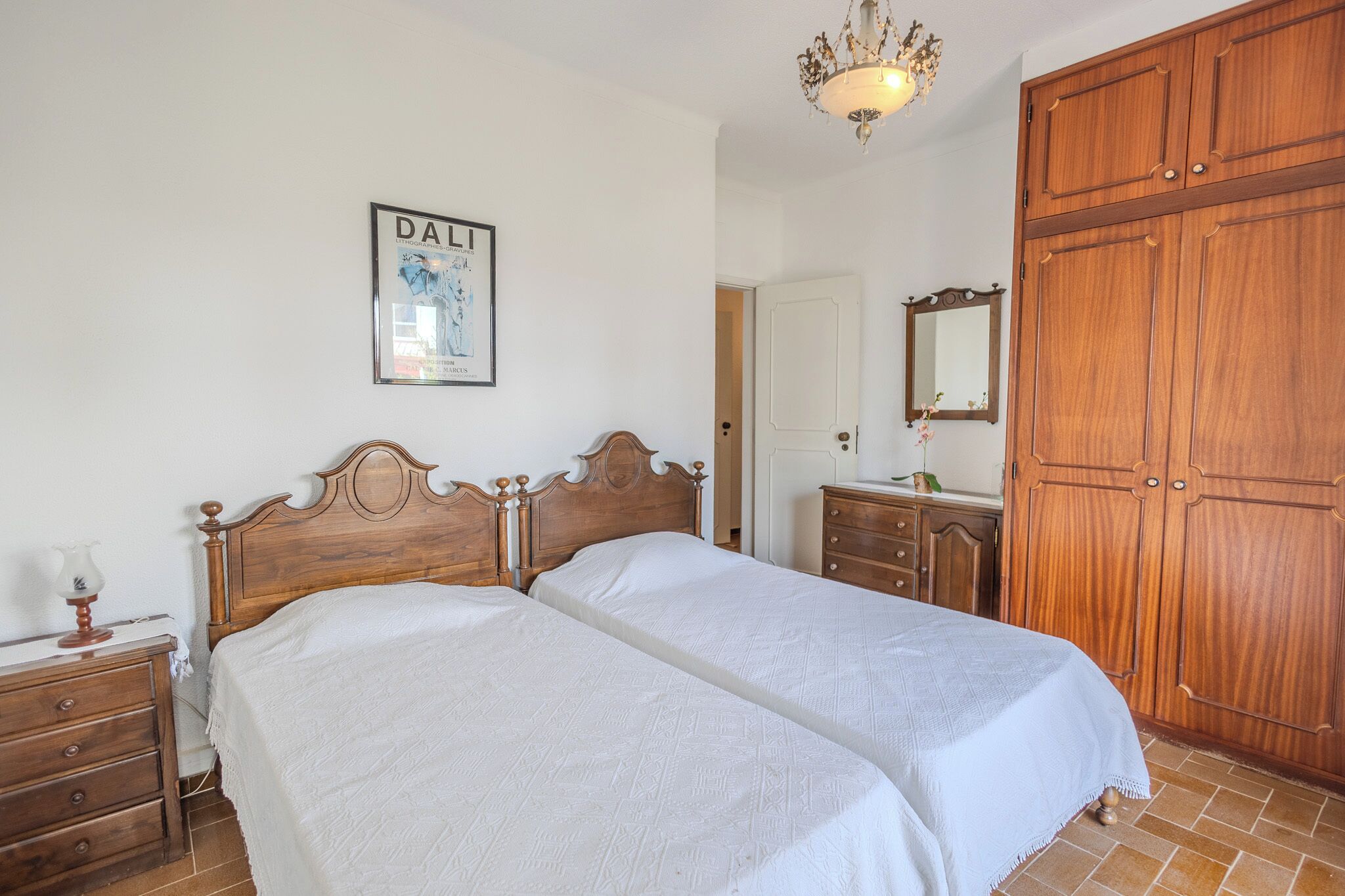 A bedroom with two beds, wooden wardrobe, a chandelier, and a framed picture on the wall.
