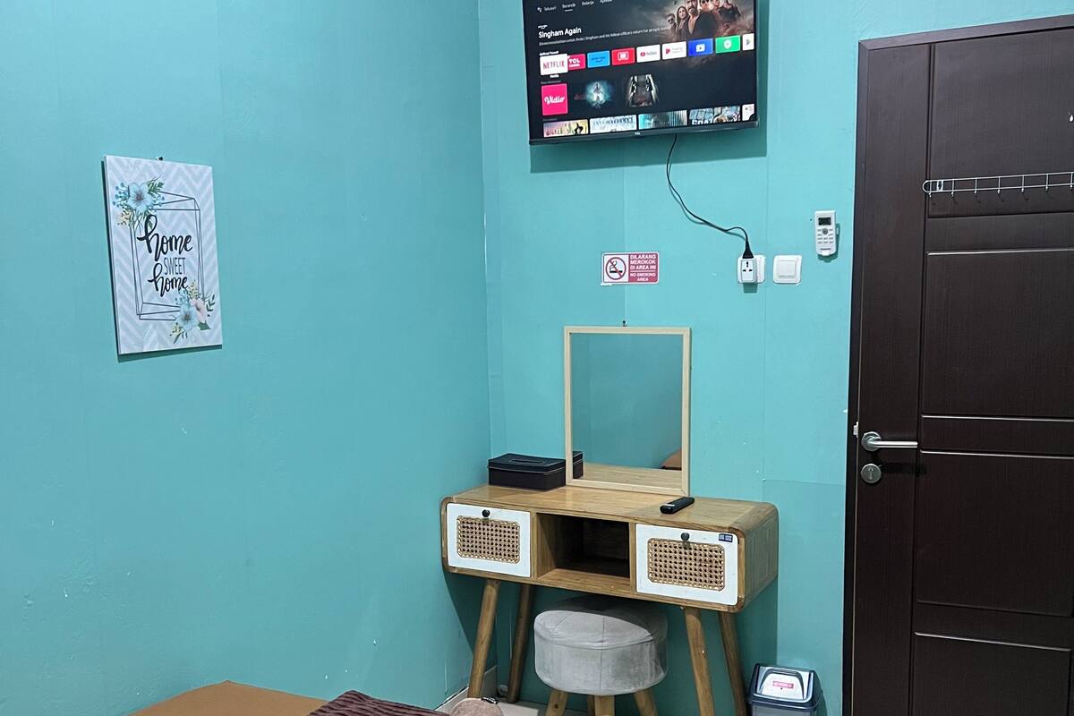 Une pièce comprenant un lit, un téléviseur fixé au mur, un bureau en bois avec un miroir, un petit tabouret et une porte.