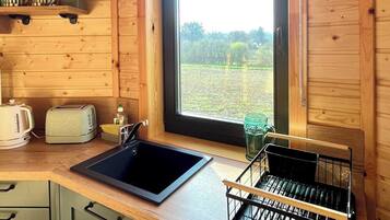 A compact kitchen with wooden walls, a window with a view of greenery, a sink, a toaster, and a dish rack.