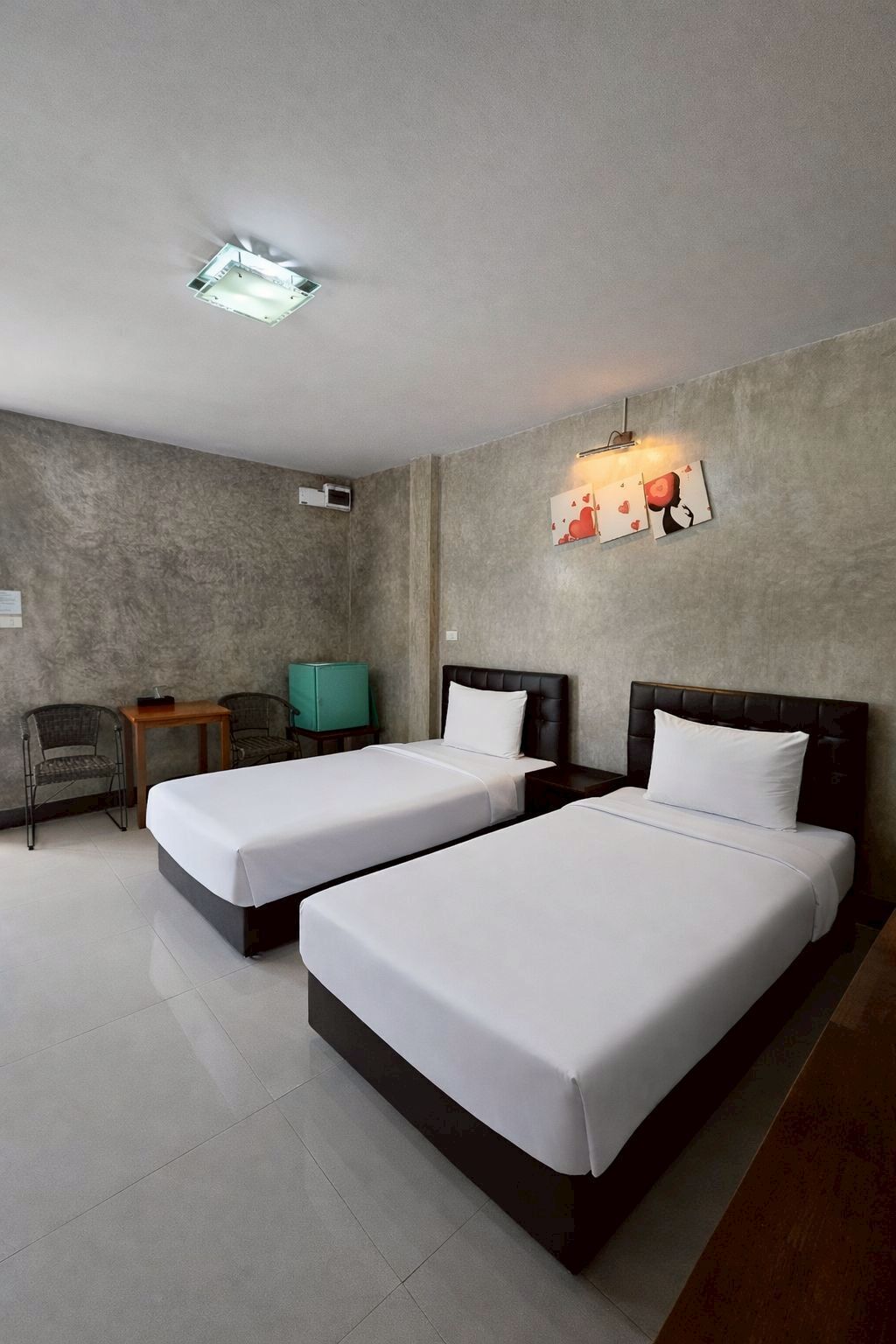 A hotel room with two beds, a small table, two chairs, a wall-mounted light, and a ceiling light.