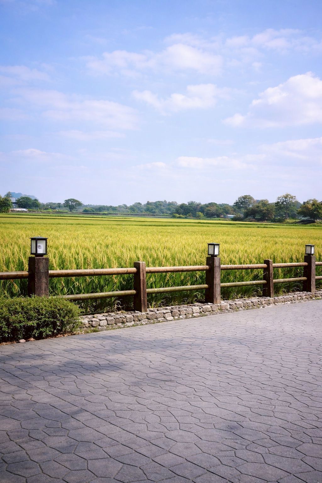 A paved walkway with a wooden fence and lanterns, leading to a green field.