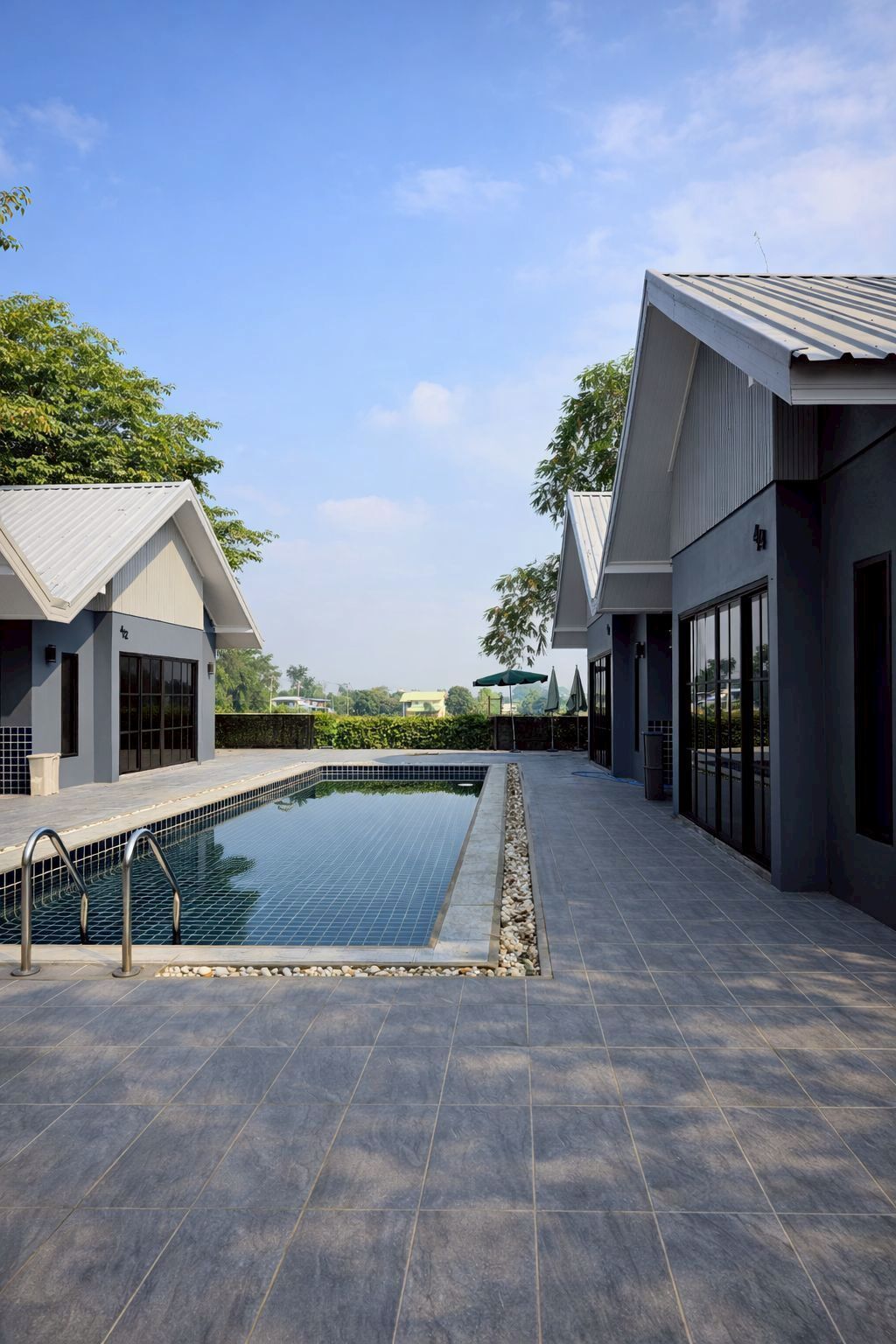 A modern outdoor swimming pool area with a clear sky, surrounded by buildings with large windows and a tiled floor.