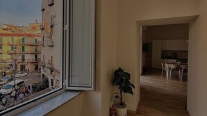A room with a large window, wooden flooring, a potted plant, and a kitchen area visible through an open door. - Cozy vacation home in the heart of the historic center of Naples (Napoli)