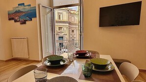 A dining area with a table set for two, a television mounted on the wall, and a view of a building through an open door. - Cozy vacation home in the heart of the historic center of Naples (Napoli)