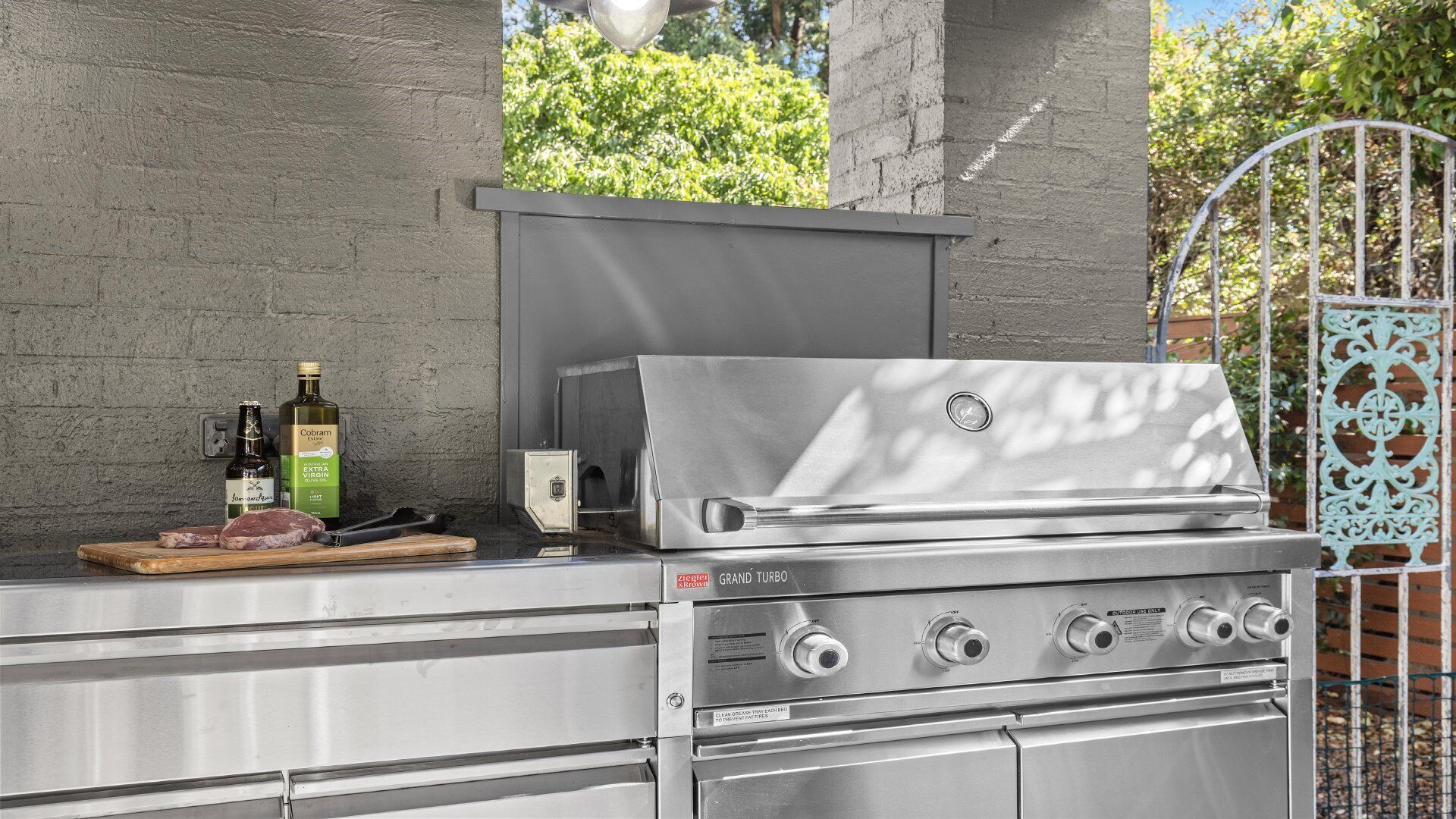 A stainless steel grill with a chimney, a bottle of wine, and a cutting board with meat.