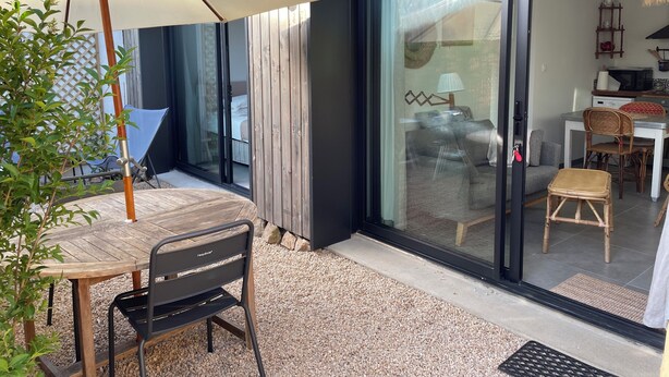 Un espace patio aménagé avec une table et des chaises, un parasol en bois et une porte-fenêtre coulissante donnant sur une pièce comprenant un lit et un coin cuisine.