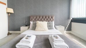 A bedroom with a tufted headboard, a bedside table with a lamp, a large fluffy pillow, and a neatly folded towel on the floor. - Incredible 3BR Battersea Flat & Spectacular View (Greater London)
