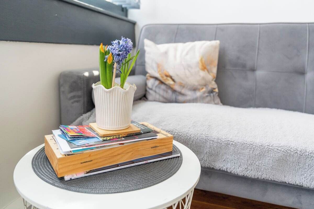 A white round table with a white vase containing flowers, a stack of books, and a magazine. A grey sofa with a patterned cushion is in the background.