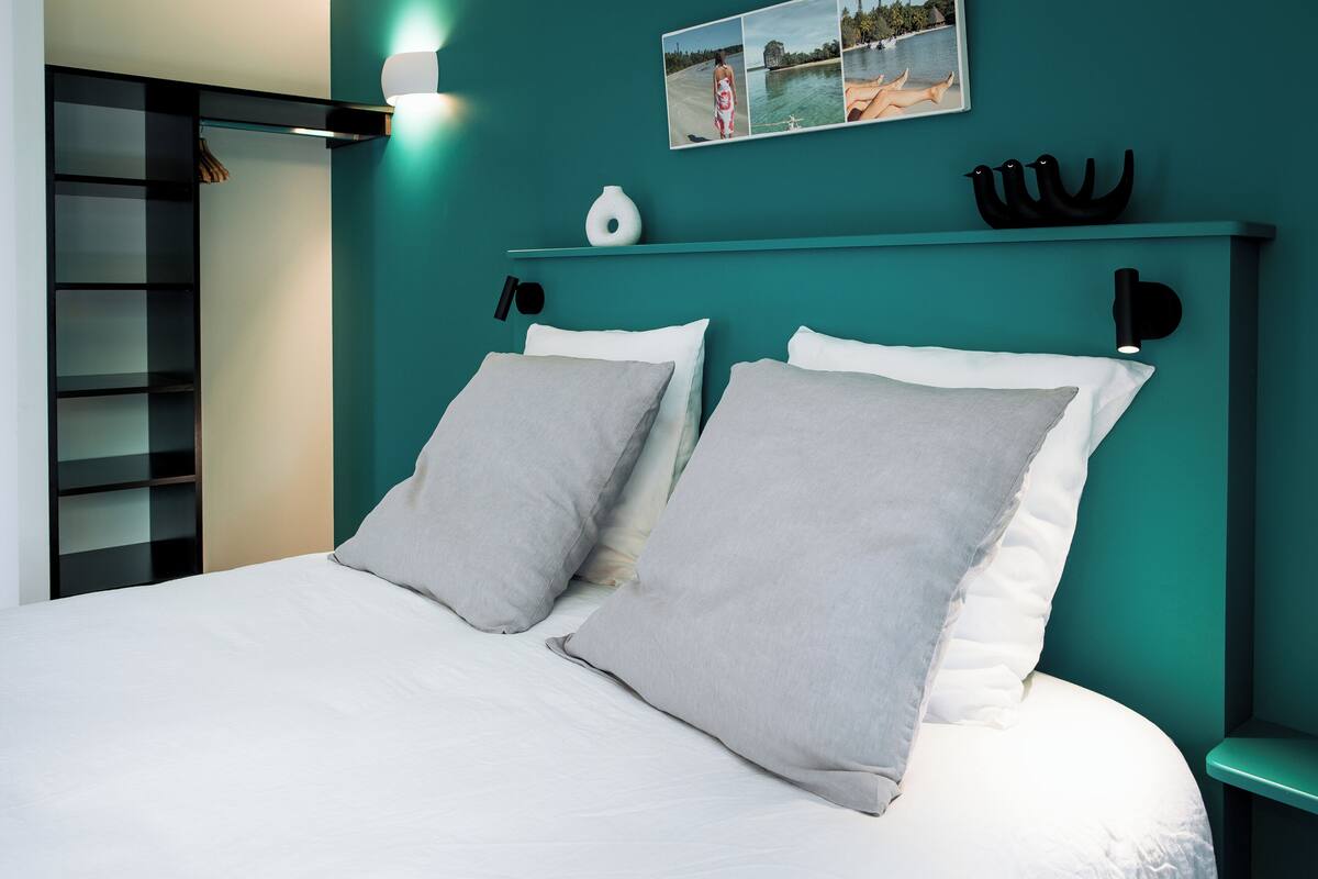 Un lit avec deux oreillers, une table de chevet, une étagère avec des objets décoratifs et une lampe fixée au mur.