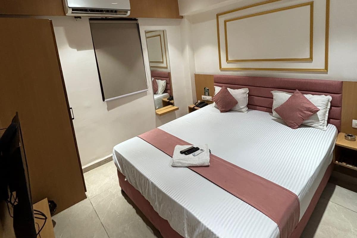 A hotel room with a large bed, a wardrobe, an air conditioning unit, a bedside table with a lamp, and a mirror on the wall.