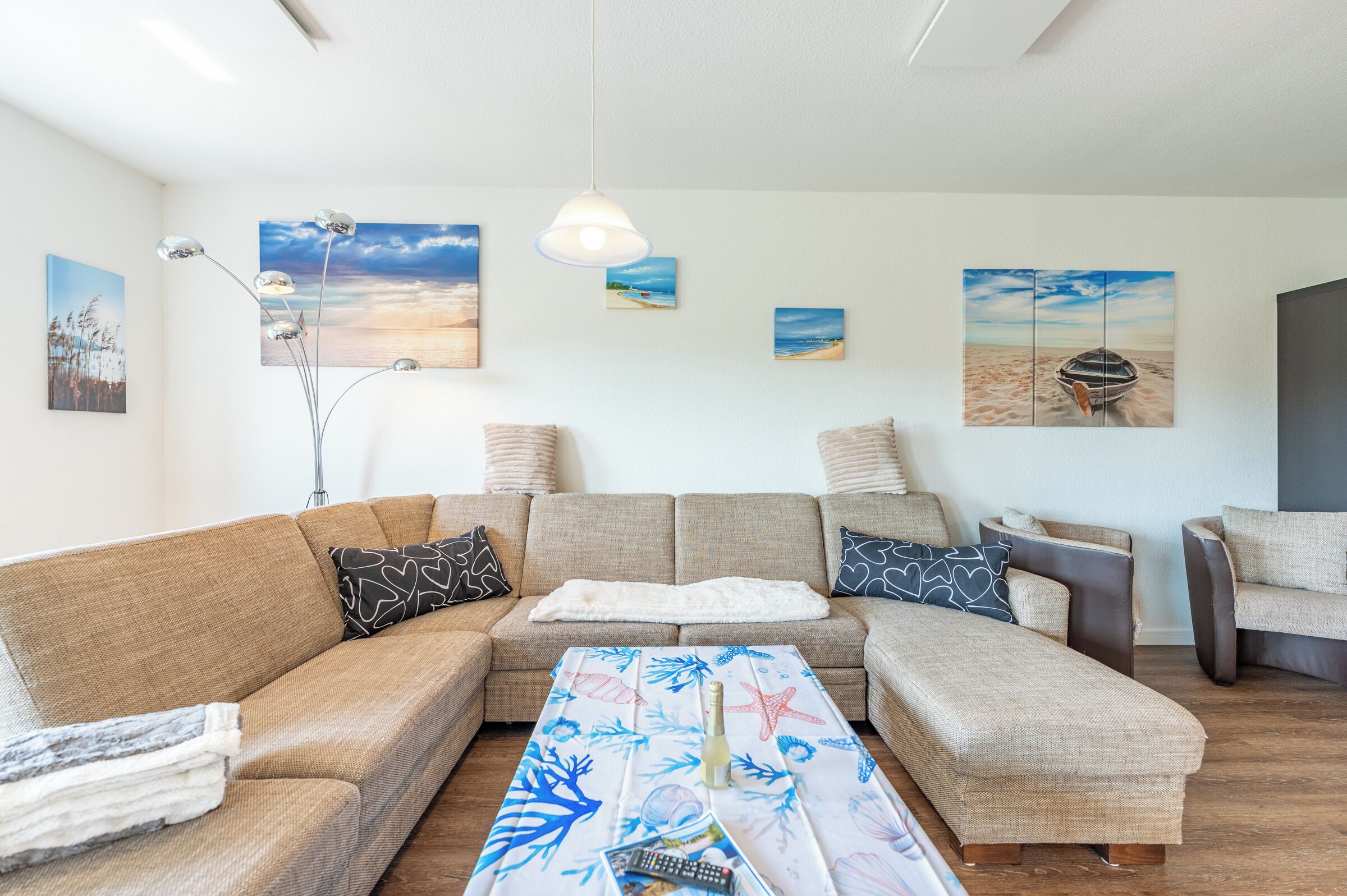 Un salon moderne avec un canapé d’angle, une table basse agrémentée d’un chemin de table sur le thème de la plage, et plusieurs tableaux accrochés au mur représentant des scènes côtières.
