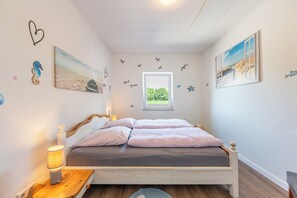 A bedroom with a bed, a nightstand, a lamp, and wall decorations including a seahorse, a beach scene, and a window with a view of greenery. - "Landgang" Vacation Rental in Husum-Schauendahl for up to 5 people (Horstedt-Feld)