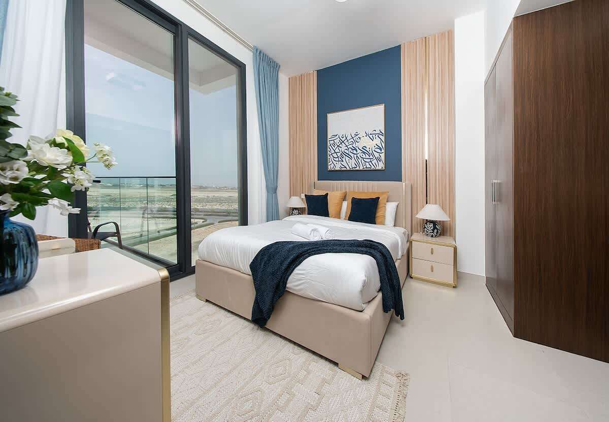 A modern bedroom with a large bed, bedside tables, a nightstand, a decorative wall piece, and a view through a large window.