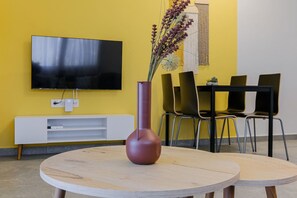 Ein Zimmer mit an der Wand montiertem Fernseher, einer weißen Unterhaltungsecke, einem runden Holztisch und einer Vase mit Blumen.