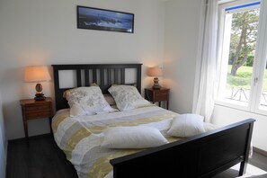 Een bed met een houten hoofdbord, twee nachtkastjes met lampen, een ingelijste foto aan de muur en een raam met uitzicht op groen.