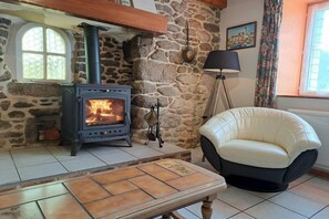 A stone fireplace with a modern stove, a cushioned chair, and a tiled floor. - Breton Stone Cottage by Sandy Beaches (Landéda)